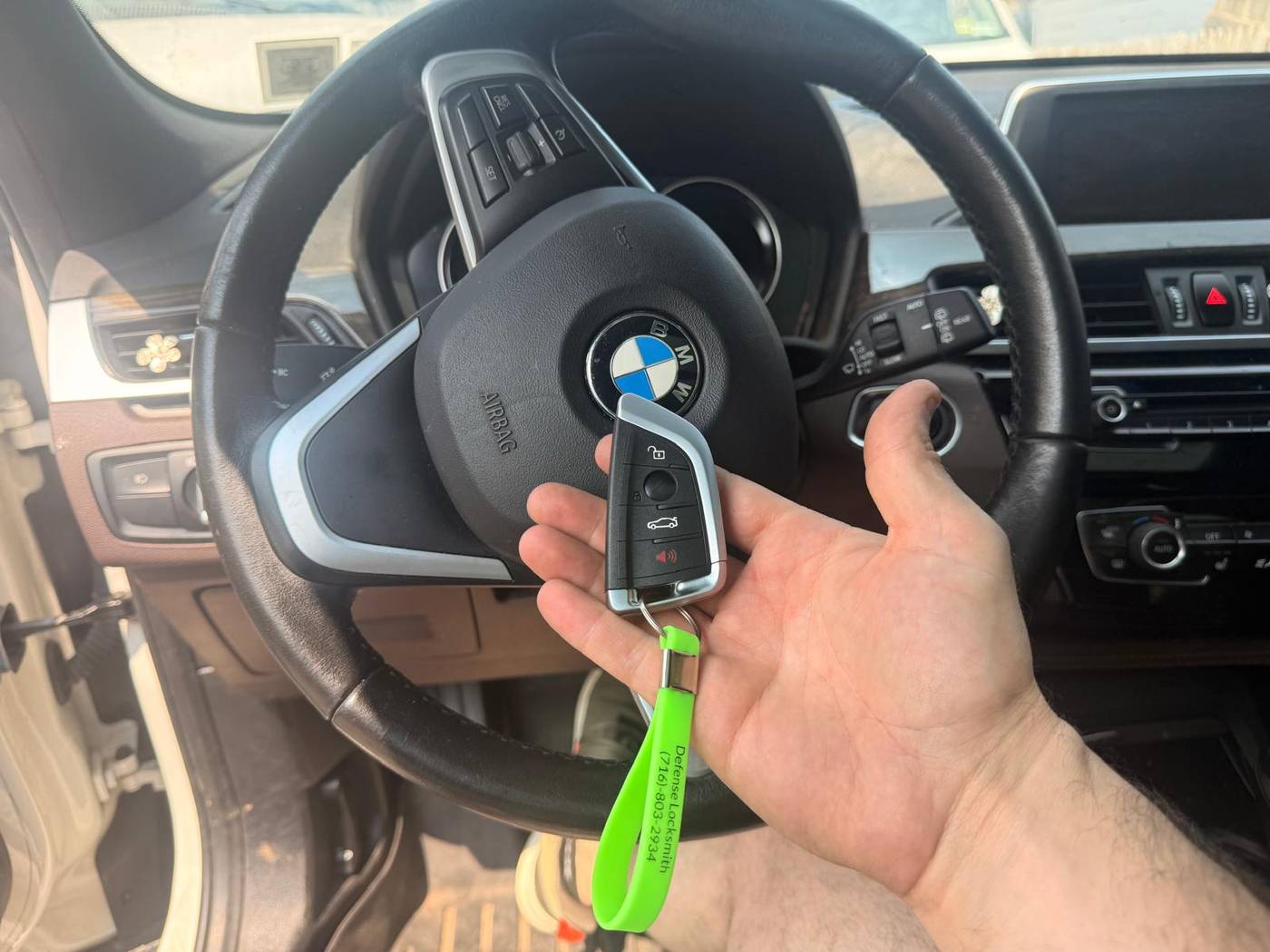 BMW X1 key made from scratch in Buffalo, NY New BMW X1 key fob with a green Defense Locksmith tag held at the steering wheel after an all-keys-lost job